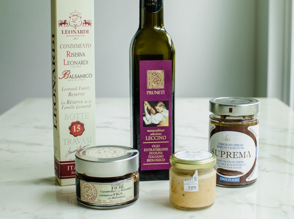 A row of Italian food products - including olive oil, balsamic vinegar, fig spread, Parmesan truffle spread, and chocolate sauce on a white marble countertop. 