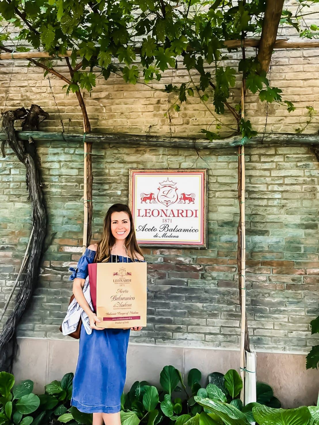 A young woman holding a shopping bag in front of a brick wall with a sign for Leonardi balsamic. 