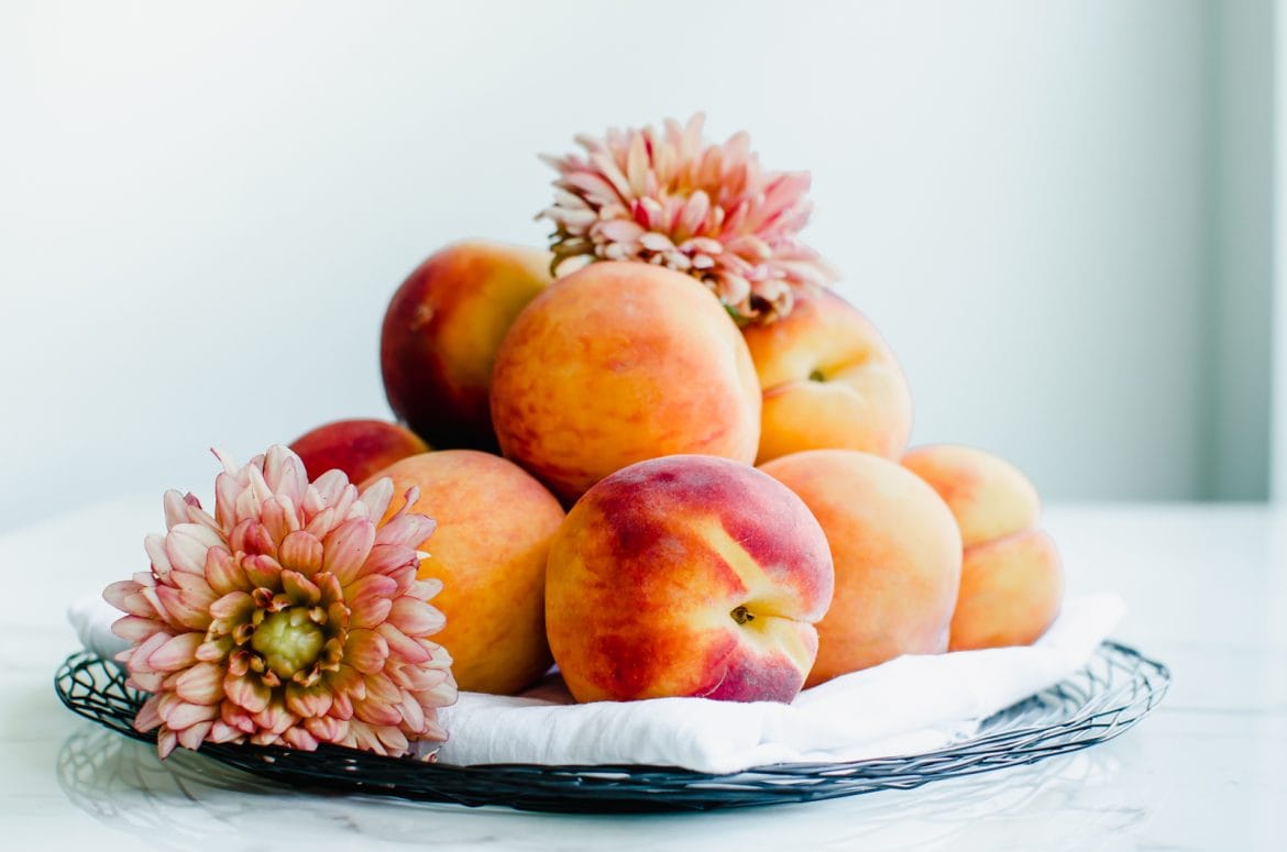 Fresh peaches piled on a wire plate with a white towel and coral dahlias sitting on the peaches.