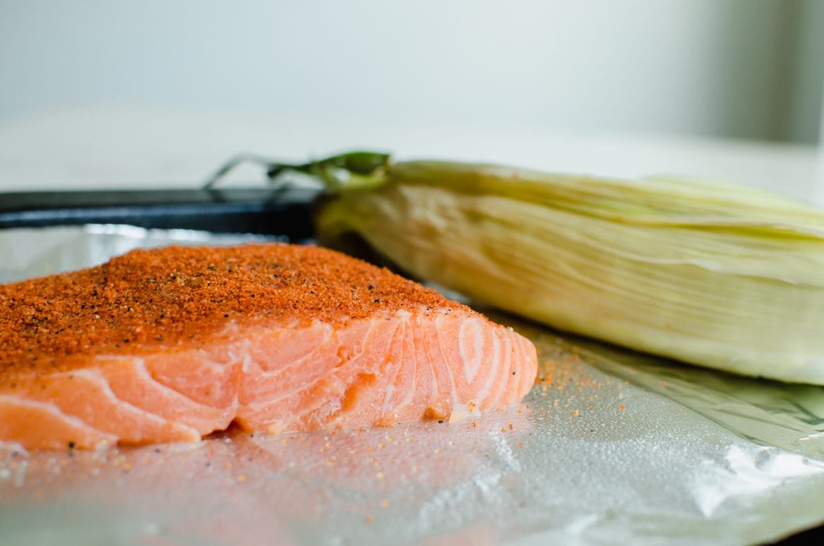 A piece of raw salmon on a sheet pan with a piece of corn in the shuck.