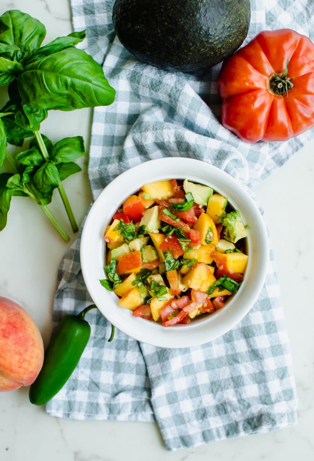 An overhead shot of a white bowl with peach salsa and produce around the sides of the bowl.