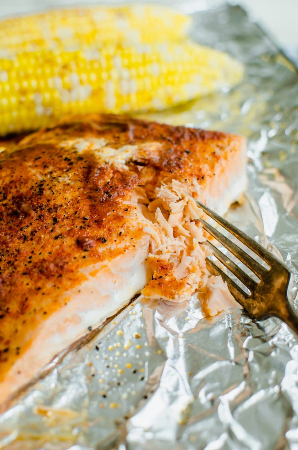 A piece of cooked BBQ salmon being flaked with a fork and a piece of roasted corn in the background.