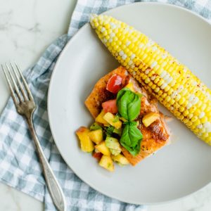 A piece of BBQ salmon on a beige plate topped with peach salsa and an ear of roasted corn on the side.