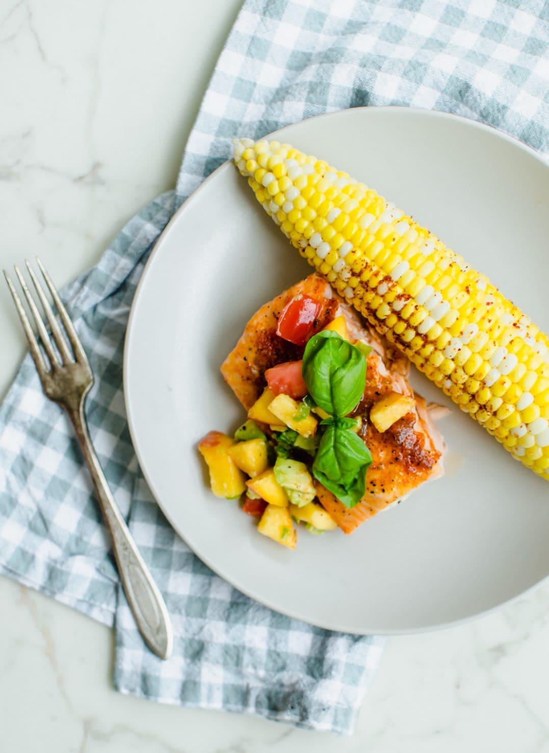 A piece of BBQ salmon on a beige plate topped with peach salsa and an ear of roasted corn on the side. 