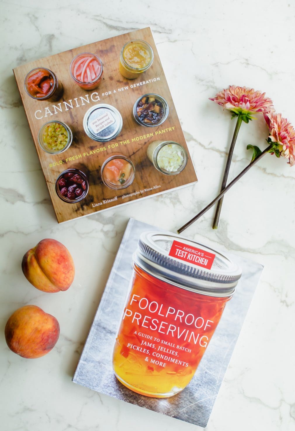 An overhead shot of two canning cookbooks with two dahlia flowers and fresh peaches on the side. 