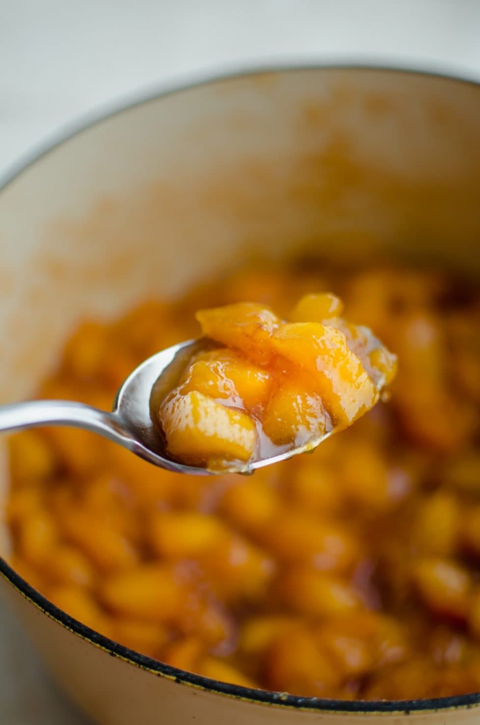 A close up shot of a spoon of peach jam over a pot of jam cooking. 