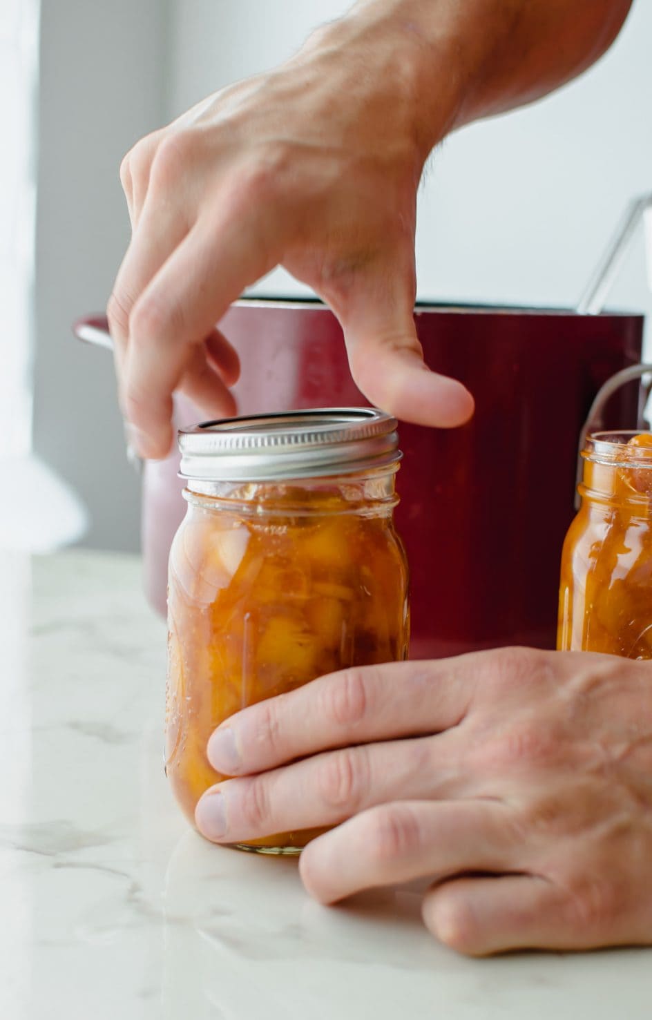 A pair of hands putting the lid on a canning jar filled with peach jam. 