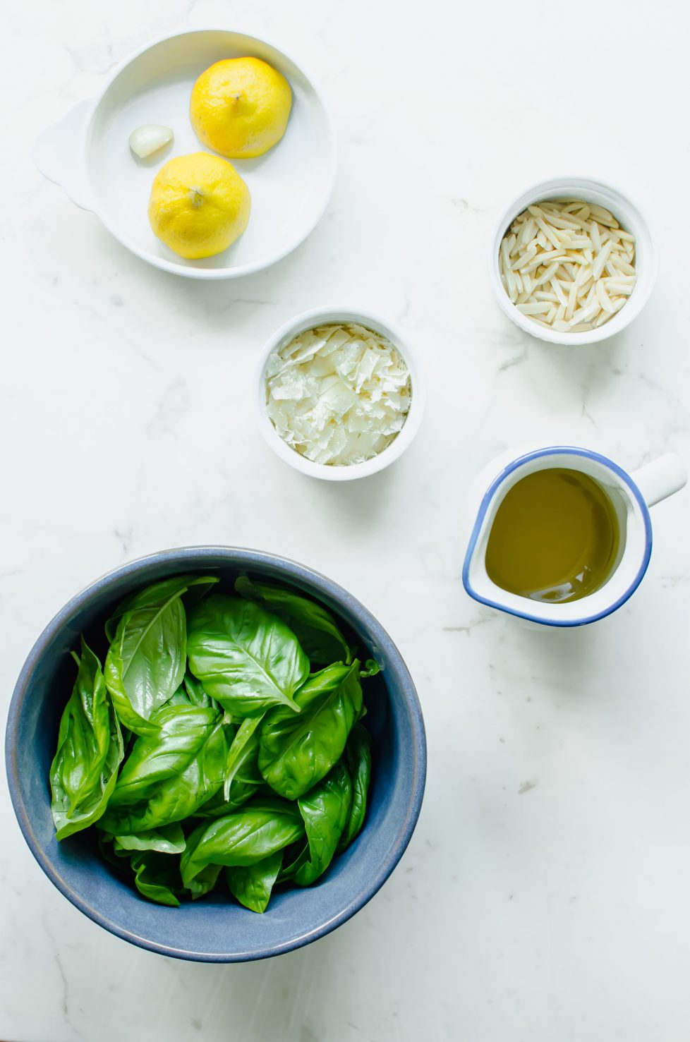 An overhead shot of small bowls of pesto ingredients on a white marble countertop.