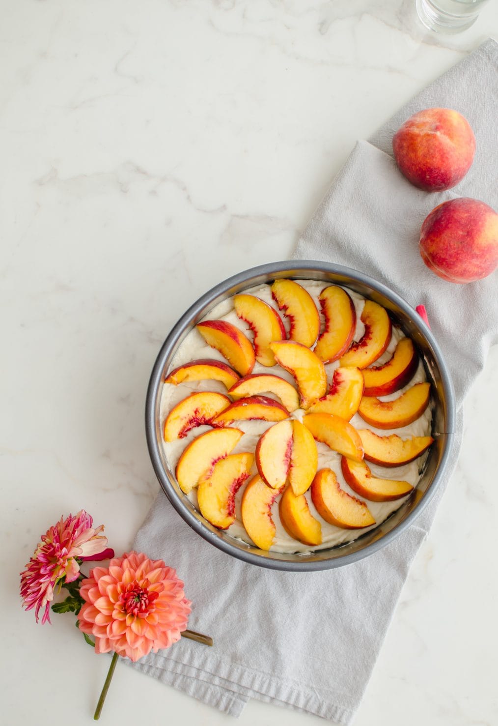 An overhead shot of an unbaked cake with yellow cake batter topped with fresh peaches on a white counter top.