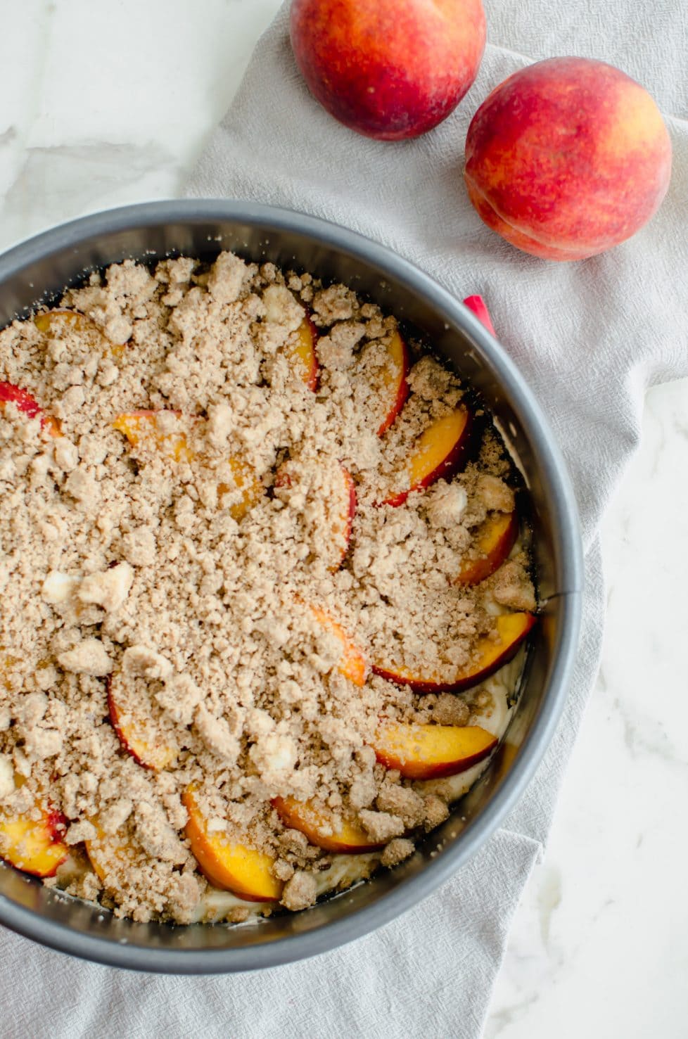 A close up shot of an unbaked peach crumb coffee cake in a springform pan on a white counter top.