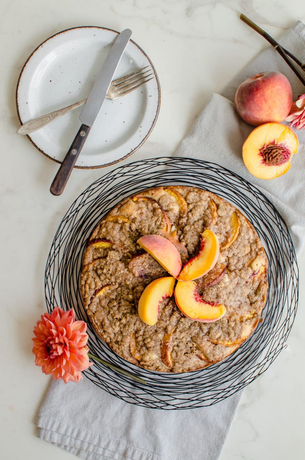 An overhead shot of a peach crumb coffee cake with sliced peaches on top, dahlias and fresh peaches on the side. 