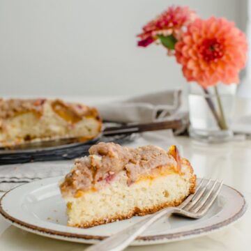 A slice of peach crumb coffee cake on a white countertop with a vase of dahlias in the background.