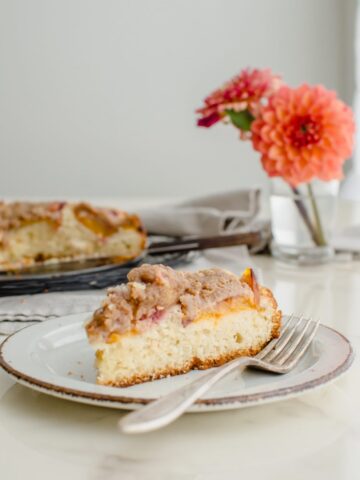 A slice of peach crumb coffee cake on a white countertop with a vase of dahlias in the background.