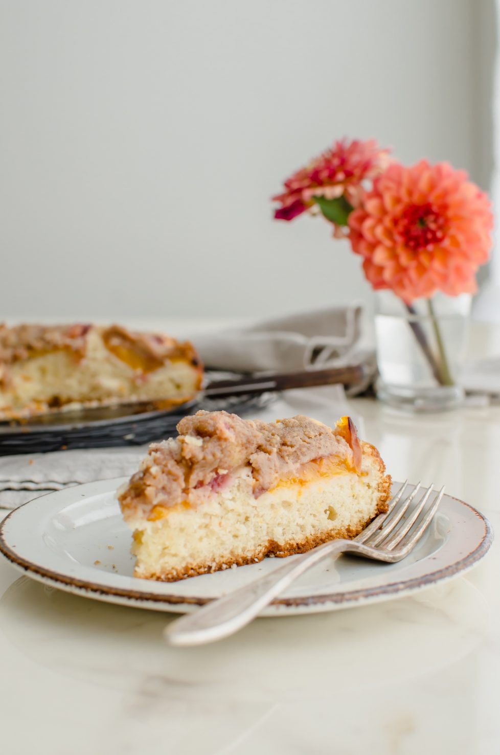 A slice of peach crumb coffee cake on a white countertop with a vase of dahlias in the background. 