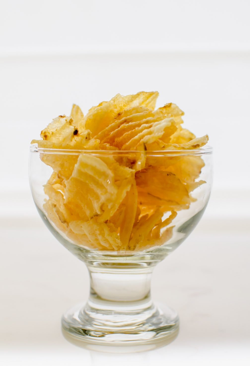 A glass bowl filled with ruffled potato chips against a white background.