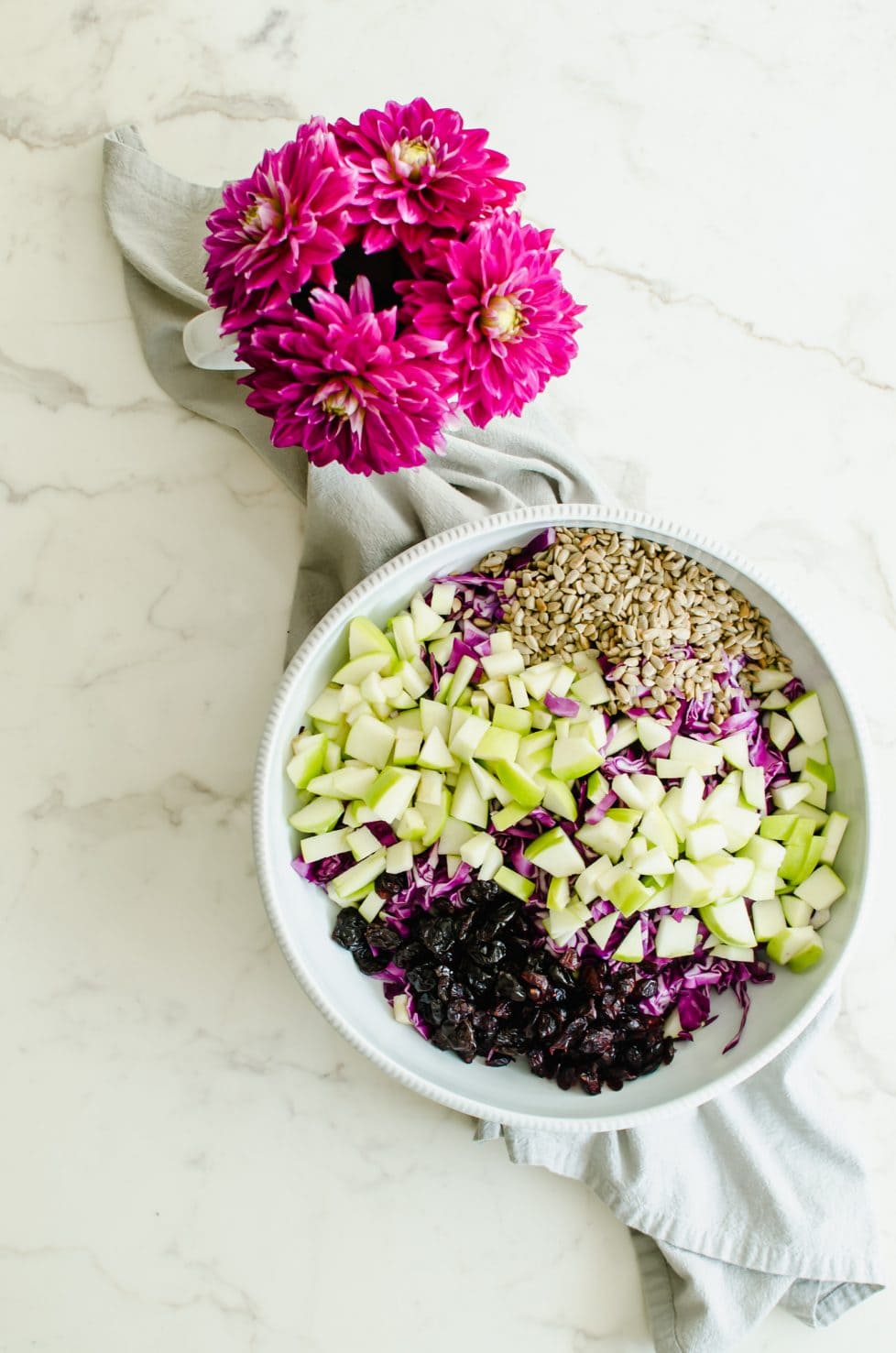 An overhead shot of shredded red cabbage, chopped green apple, sunflower seeds, and dried cherries in a large white bowl.