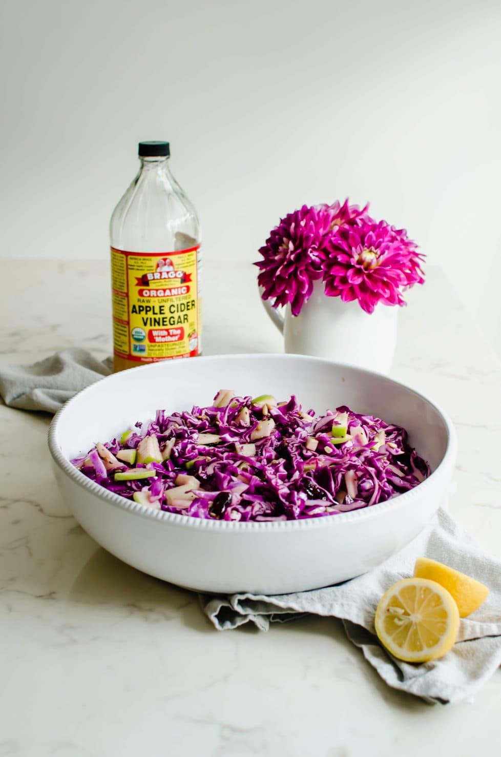 A large white bowl with red cabbage apple slaw, a bottle of cider vinegar, and a bouquet of pink dahlias.