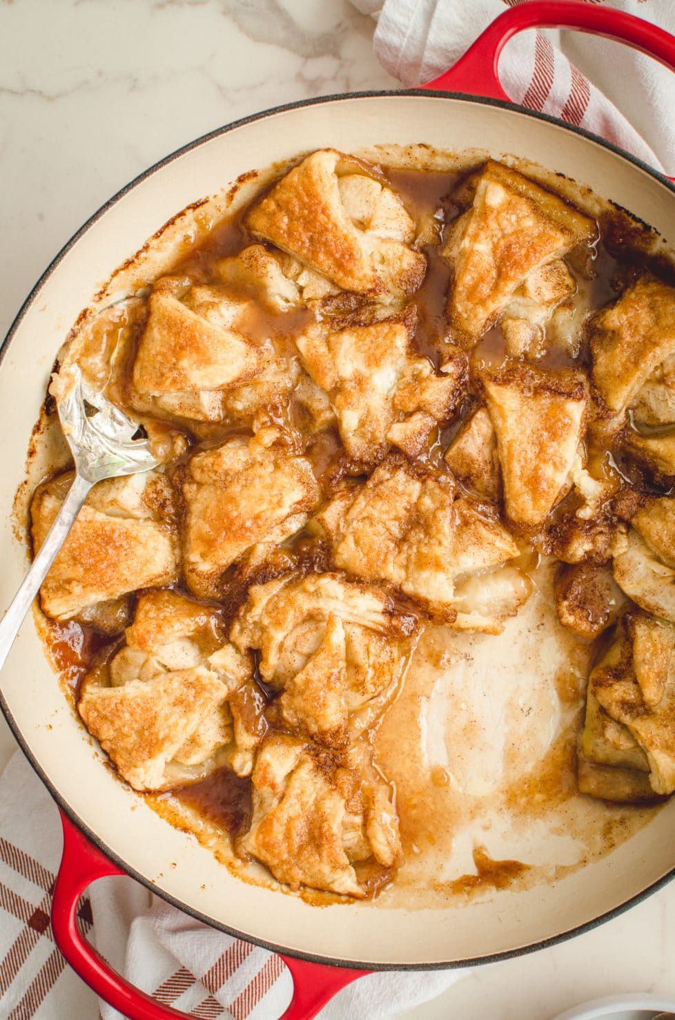 An overhead shot of apple dumplings in a red cast iron braised on a white counter top. 