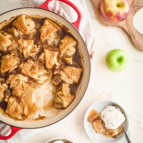 An overhead shot of apple dumplings in a red cast iron braised on a white counter top.