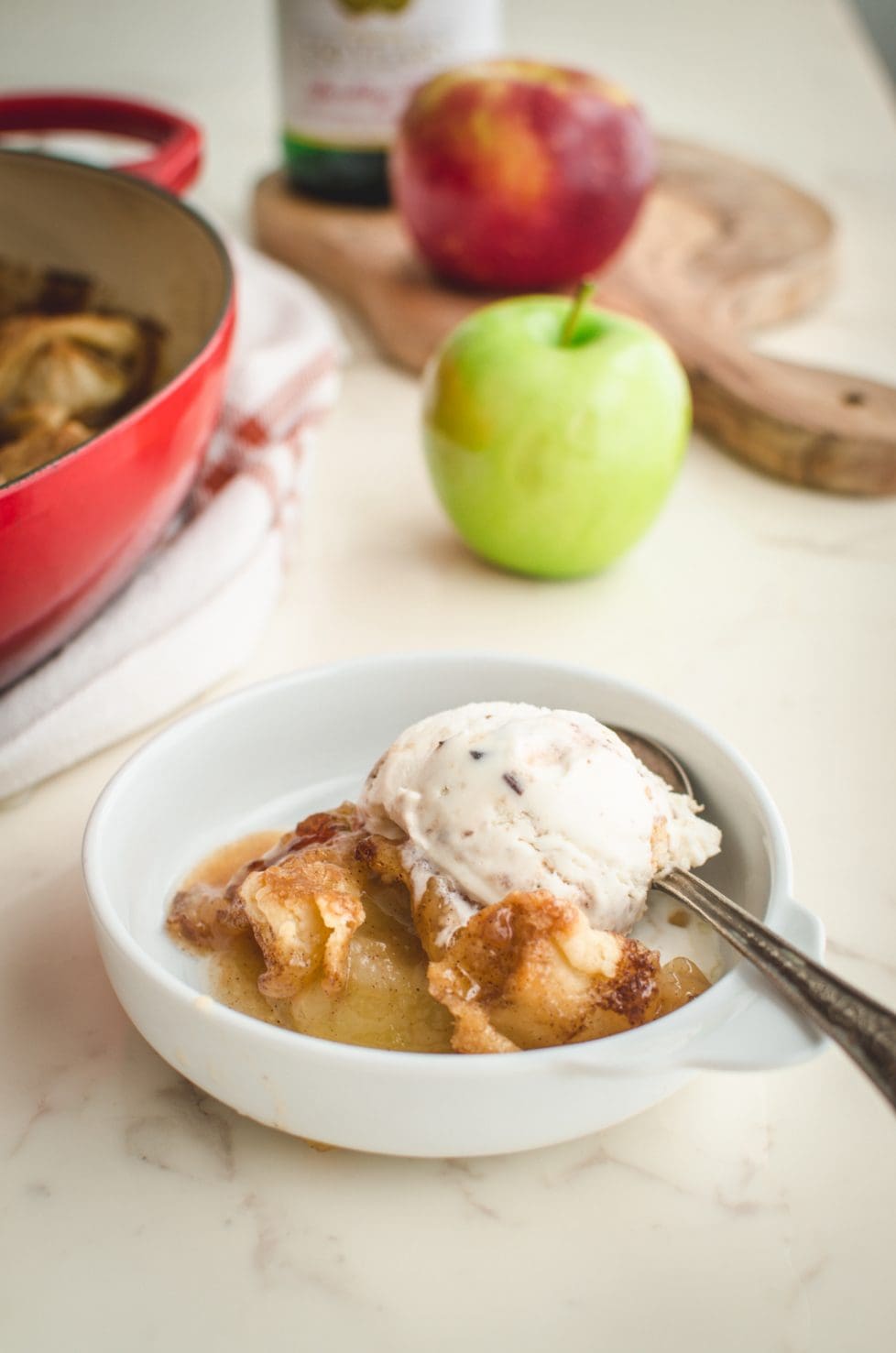 A white bowl with an apple dumpling topped with a scoop of ice cream on a white marble countertop. 