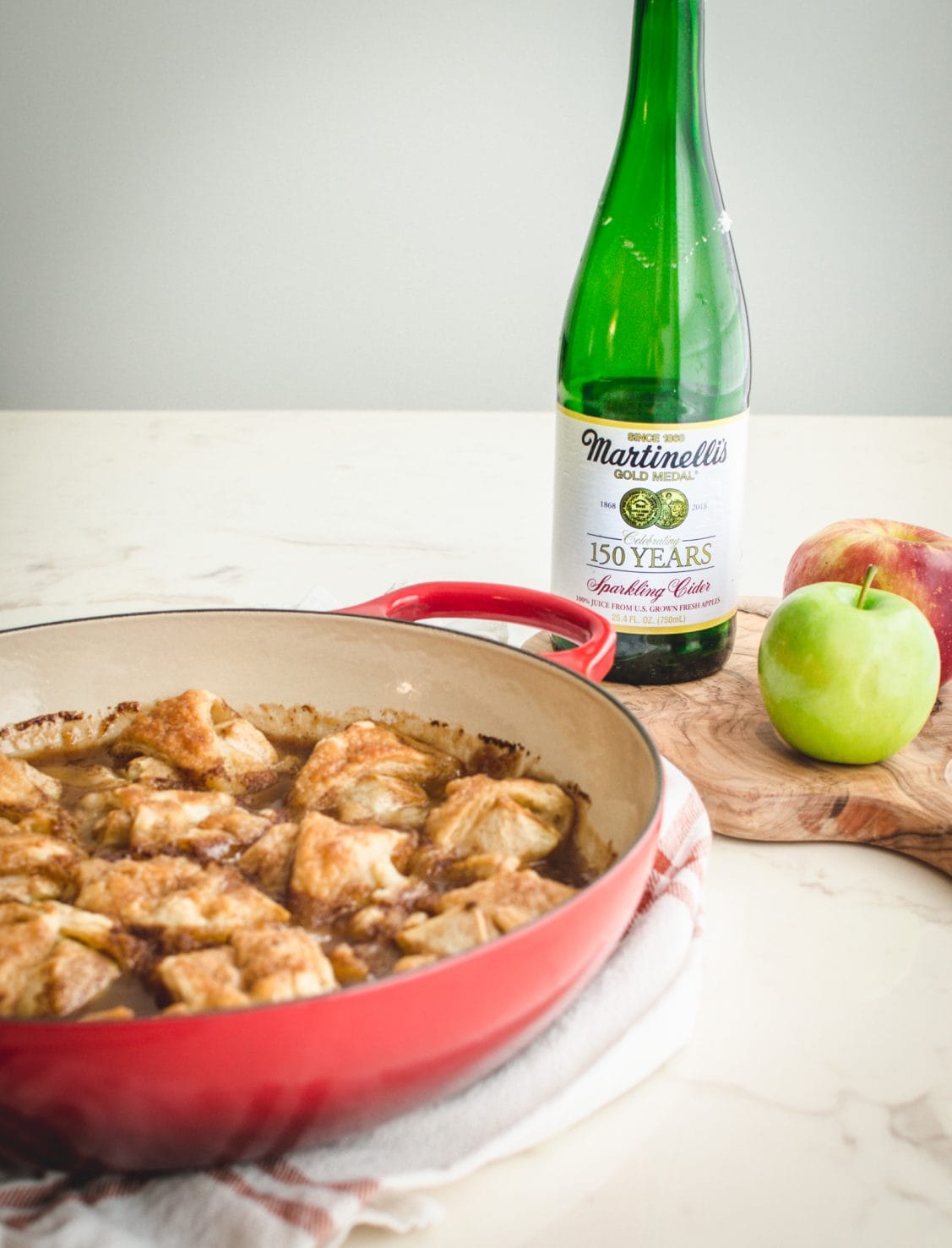 A red cast iron braised with apple dumplings with a bottle of sparkling cider in the background. 