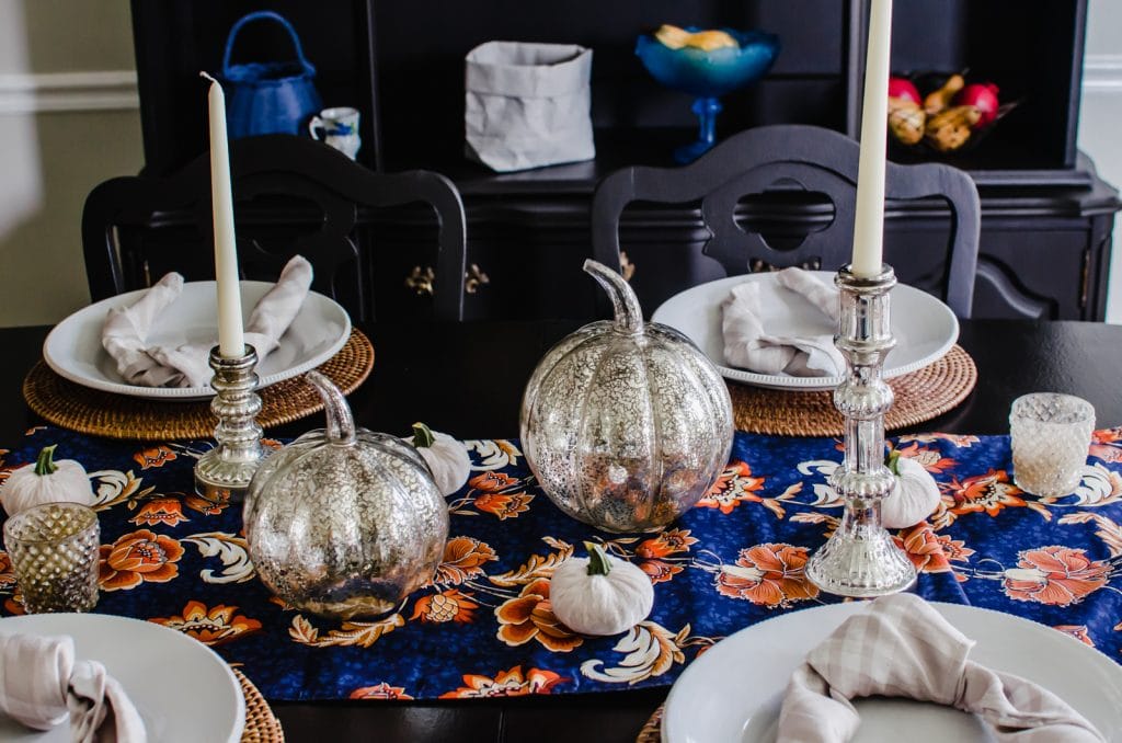 A fall table decor setting with mercury glass pumpkins, candlesticks, and a navy and orange table runner. 