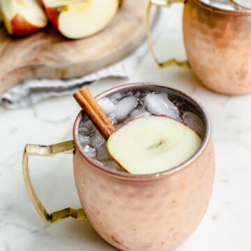 A moscow mule mug with a sparkling apple cider mocktail inside and a garnish of apple sliced and cinnamon stick.