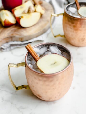 A moscow mule mug with a sparkling apple cider mocktail inside and a garnish of apple sliced and cinnamon stick.