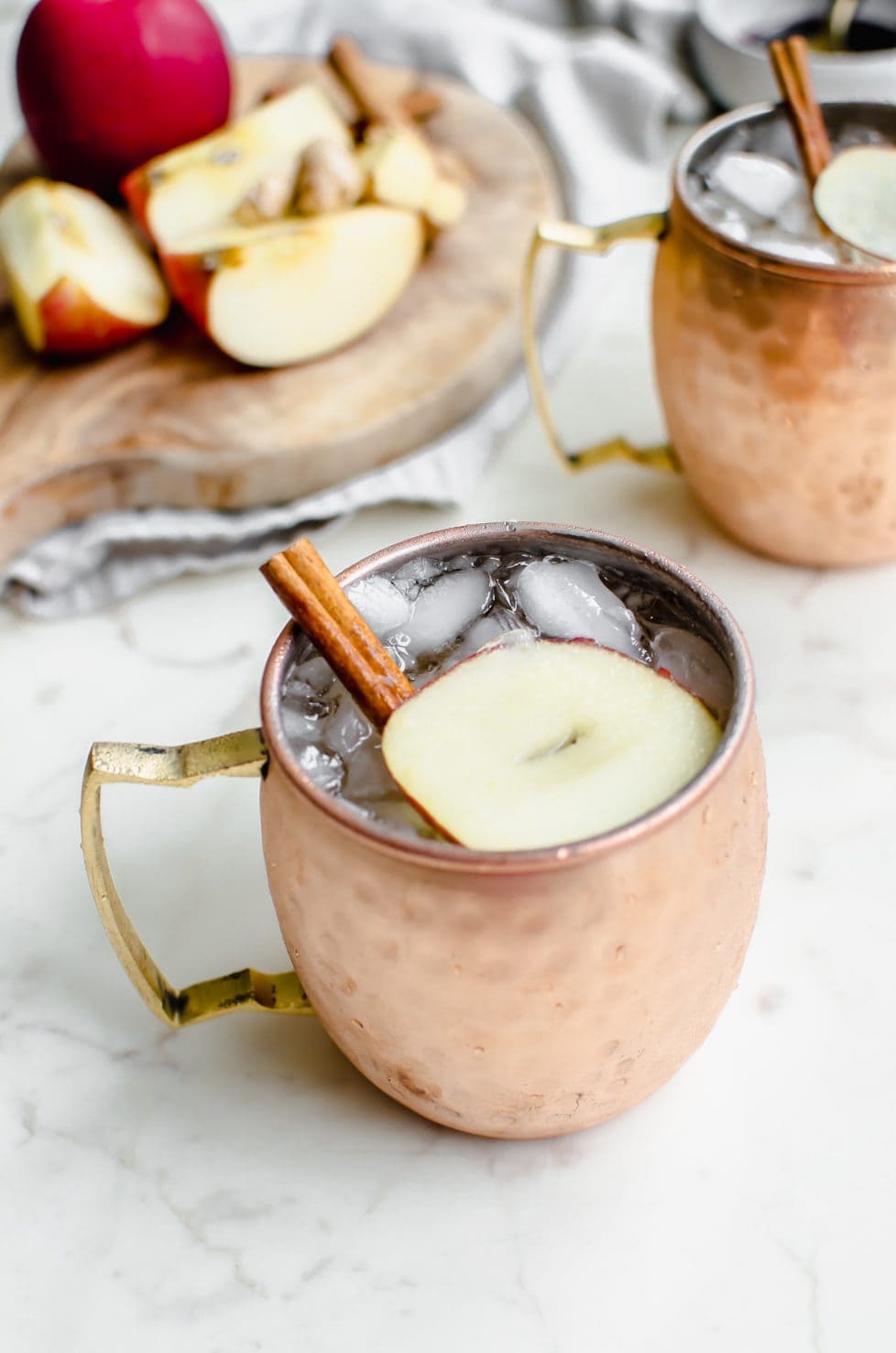 A moscow mule mug with a sparkling apple cider mocktail inside and a garnish of apple sliced and cinnamon stick.