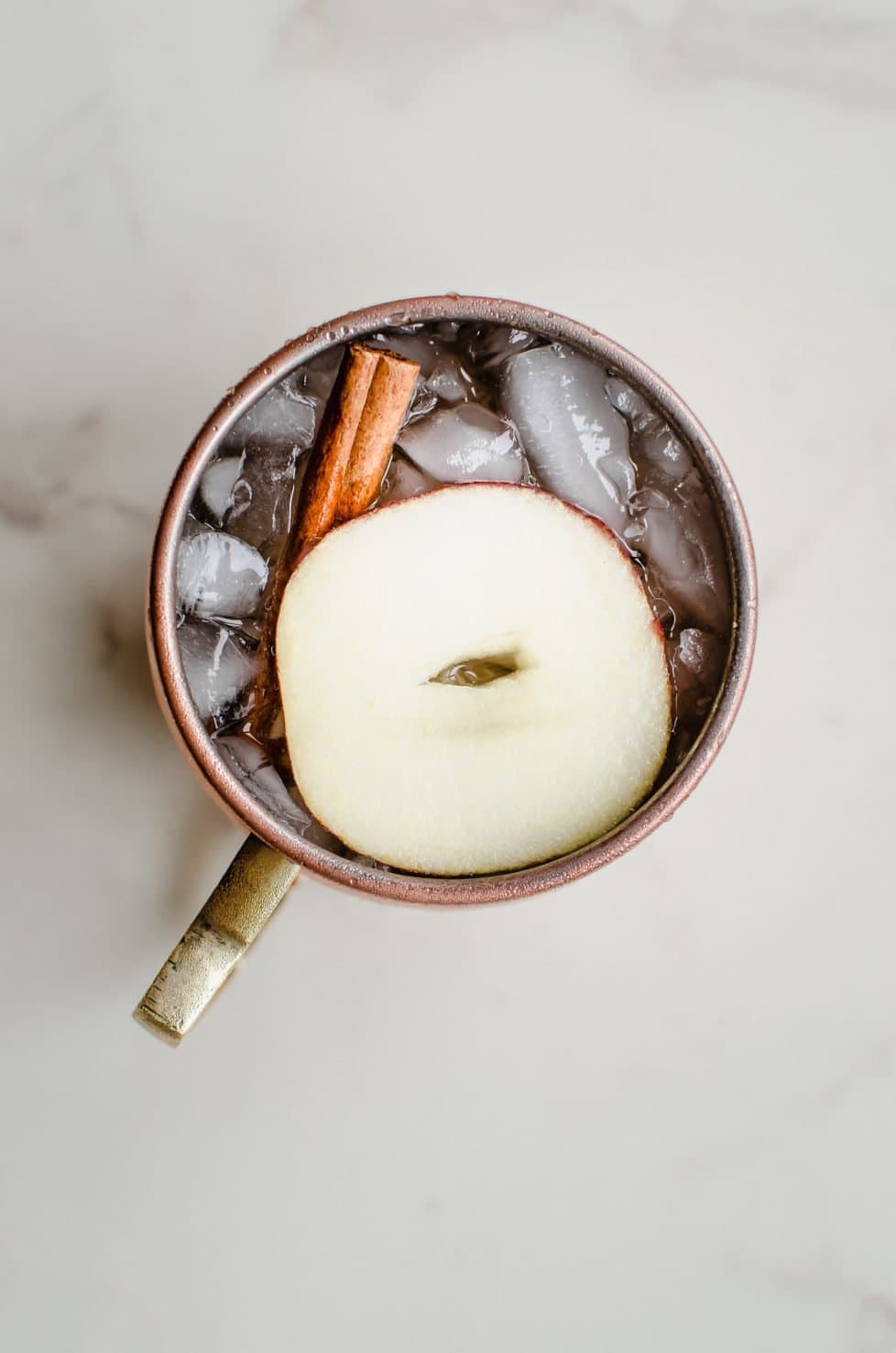 A moscow mule mug with a sparkling apple cider mocktail inside and a garnish of apple sliced and cinnamon stick. 