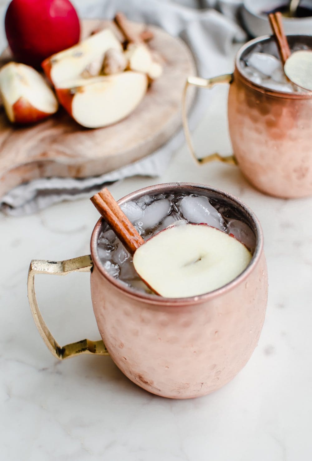 A moscow mule mug with a sparkling apple cider mocktail inside and a garnish of apple sliced and cinnamon stick. 