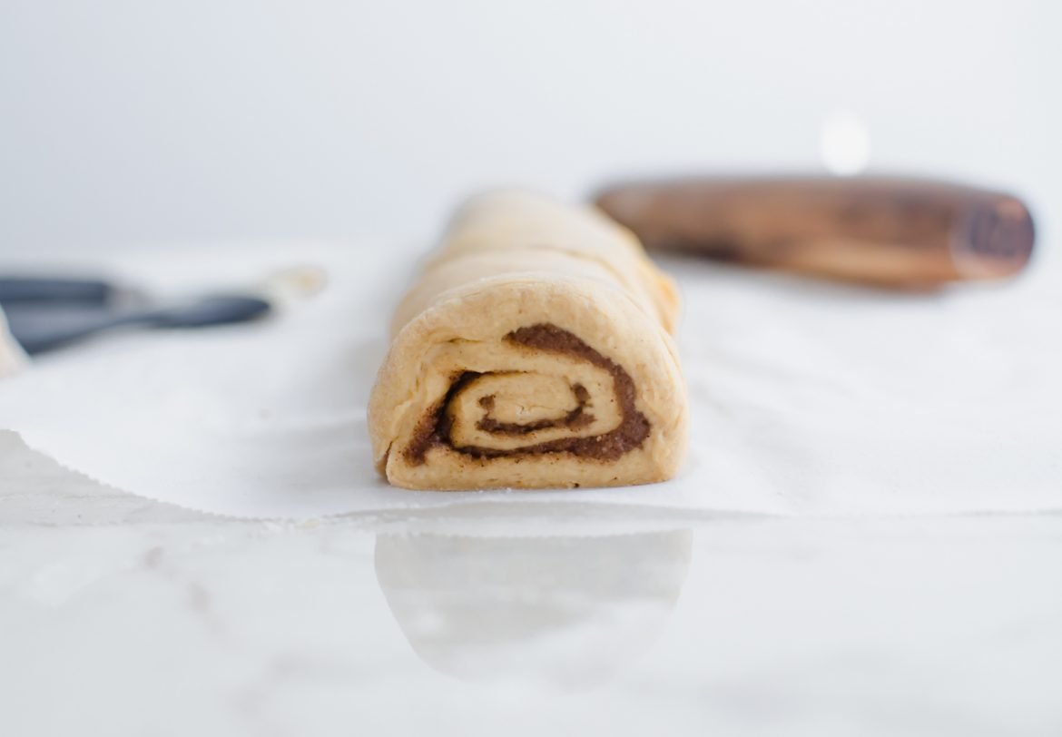 A straight-on shot of a log of rolled cinnamon roll dough with one cut taken out. 