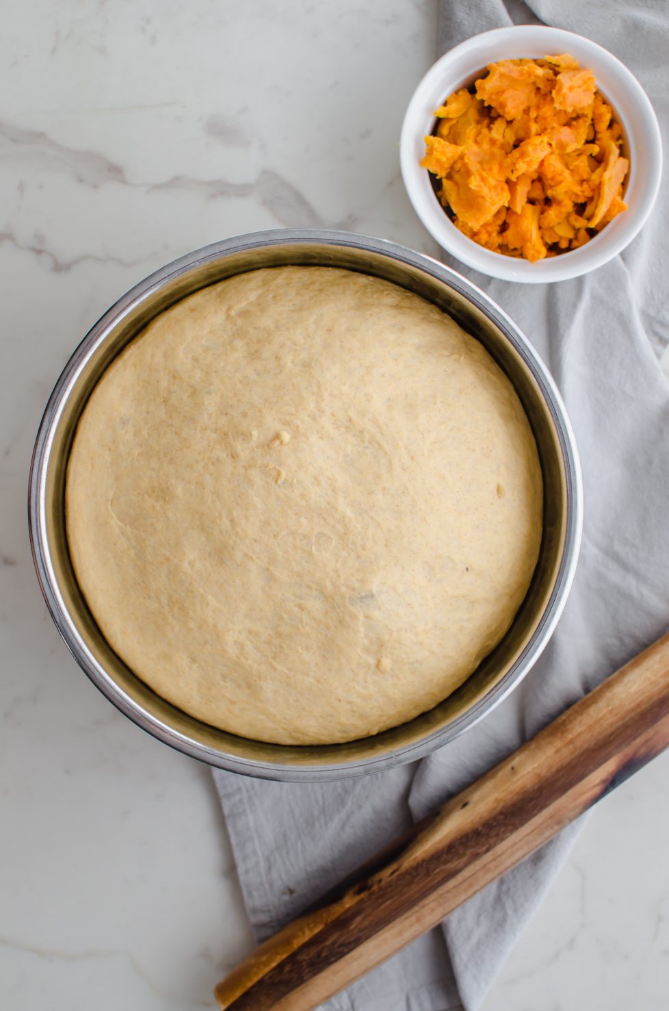 A bowl of risen sweet potato cinnamon roll dough with a rolling pin and grey towel on the side. 