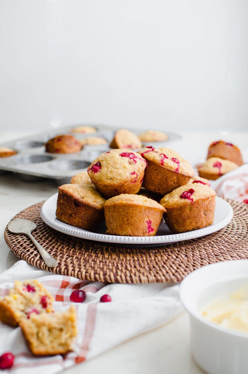 A white plate of cranberry cornbread muffins with a muffin pan in the background and bowl of honey butter on the side. 