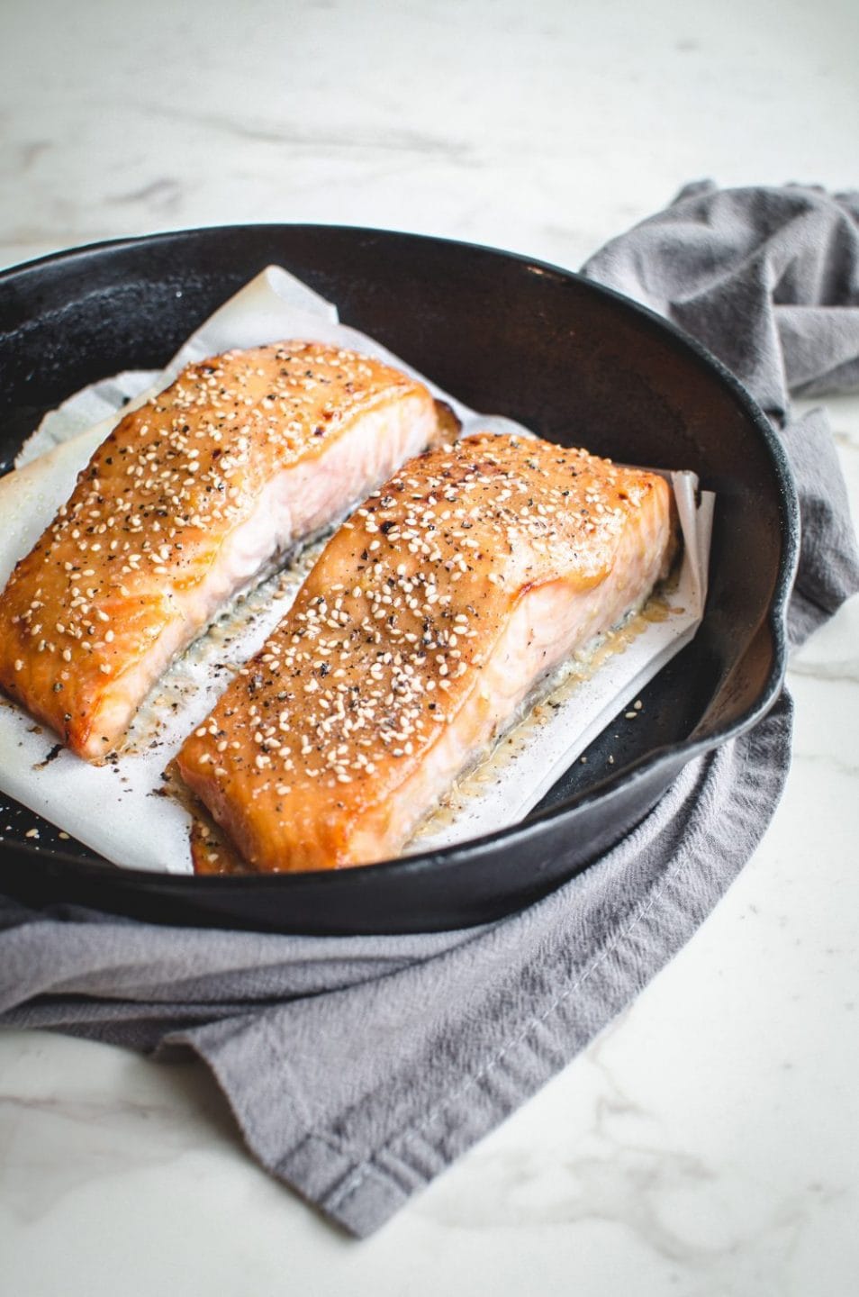 A side shot of two salmon fillets in a cast iron skillet on top of a piece of parchment paper. 