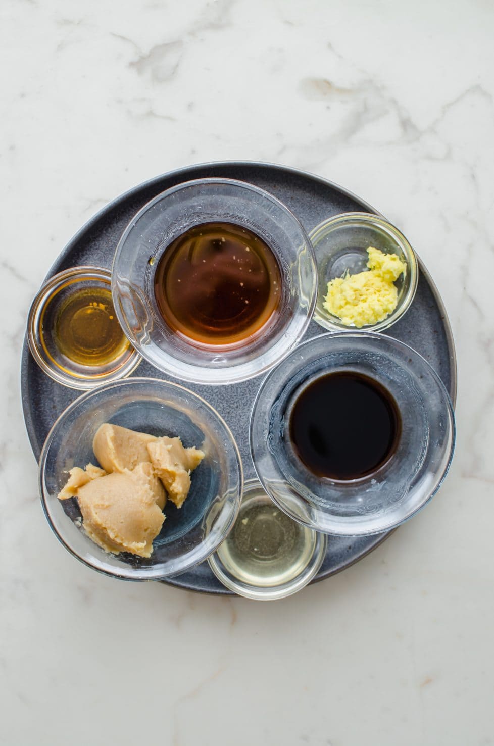 An overhead shot of prep bowls on a gray plate containing ingredients for a miso marinade.