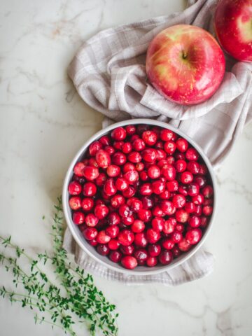 An overhead shot of a gray bowl with fresh cranberries, two apples and sprigs of thyme on the side.