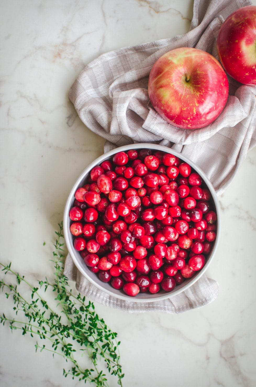 An overhead shot of a gray bowl with fresh cranberries, two apples and sprigs of thyme on the side. 