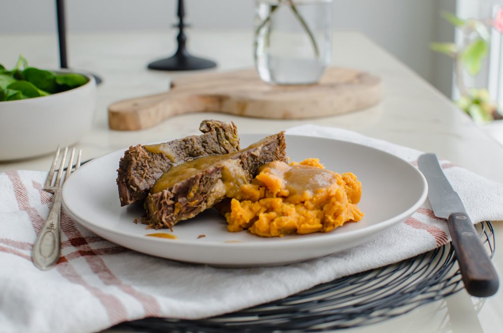 A side shot of a stone plate with beef pot roast, sweet potatoes, and cider gravy on top against a white marble background. 