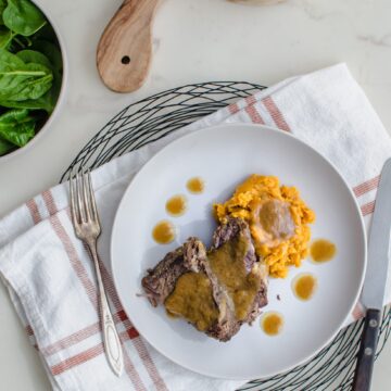 An overhead shot of a stone plate with beef pot roast, sweet potatoes, and cider gravy. A bowl of raw spinach is on the side.