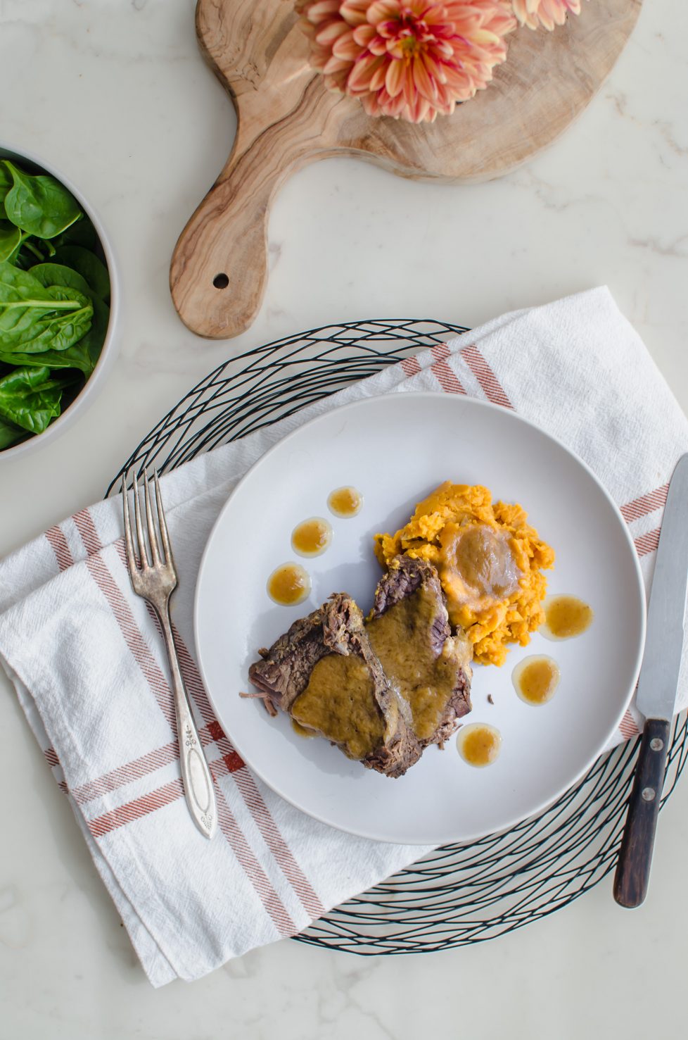 An overhead shot of a stone plate with beef pot roast, sweet potatoes, and cider gravy. A bowl of raw spinach is on the side. 