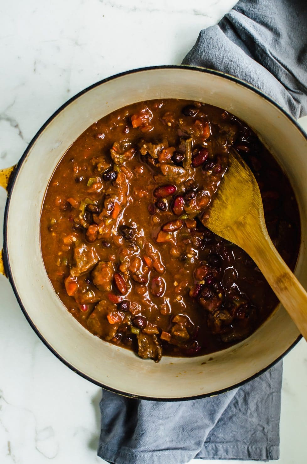 An overhead shot of a yellow Dutch oven filled with Texas Steak Fajita Chili and a wooden spoon.