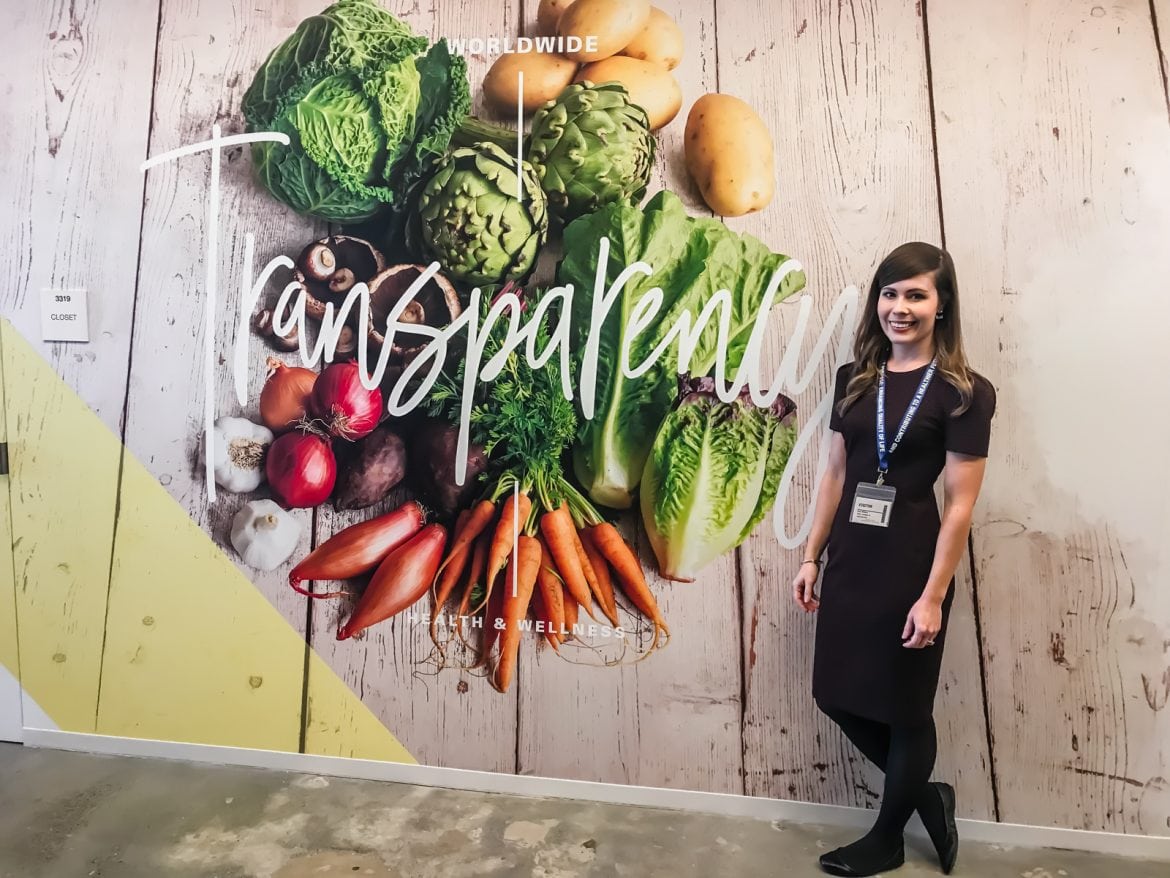 A young woman standing in front of a wall with a picture of produce and the word Transparency.