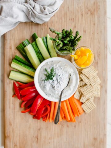 A cutting board with colorful veggies surrounding a white bowl with creamy Parmesan herb dip.