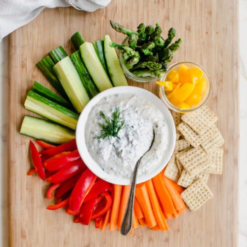 A cutting board with colorful veggies surrounding a white bowl with creamy Parmesan herb dip.