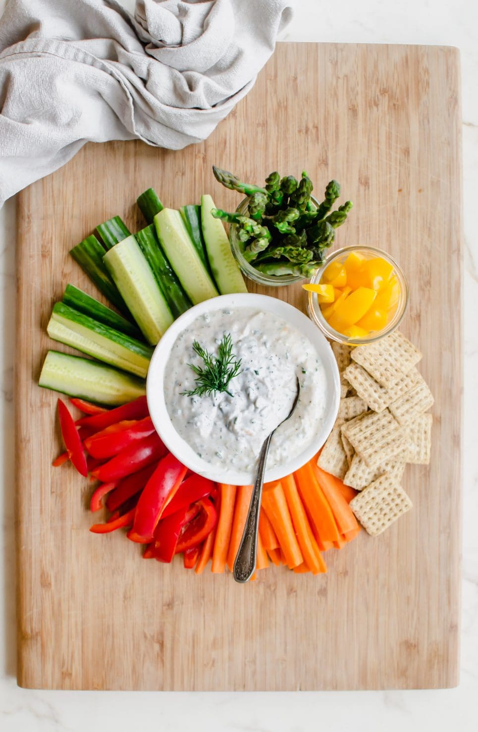 A cutting board with colorful veggies surrounding a white bowl with creamy Parmesan herb dip.