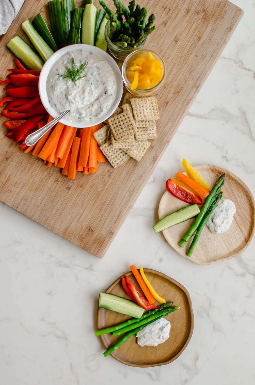 A cutting board with colorful veggies surrounding a white bowl with creamy Parmesan herb dip. 