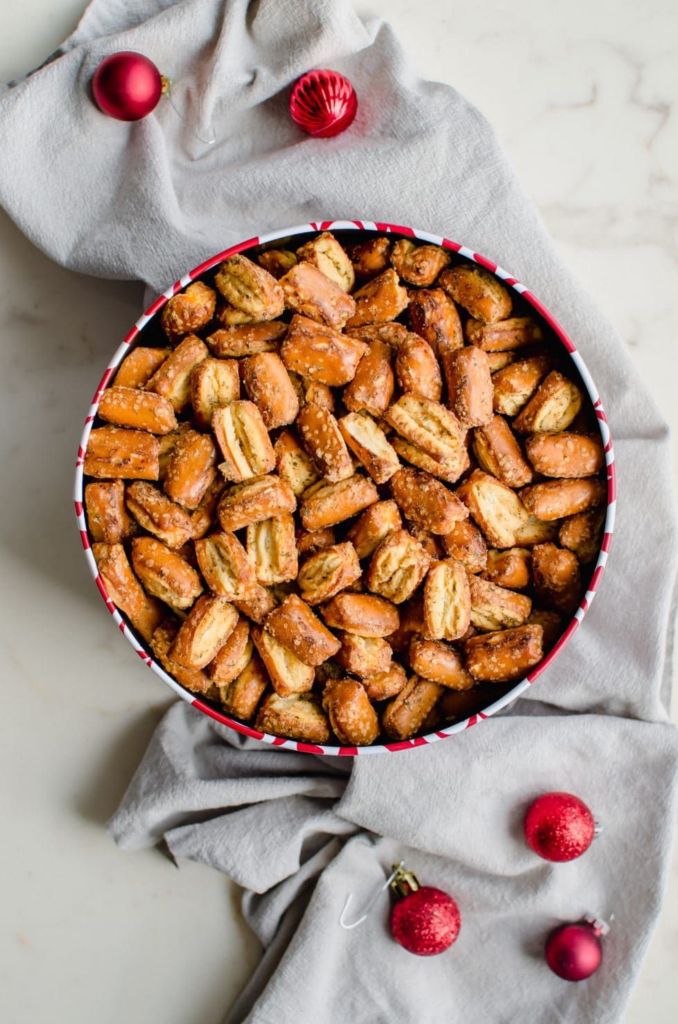 A Christmas treat tin filled with Ranch pretzel mix sitting on a grey dish towel. 