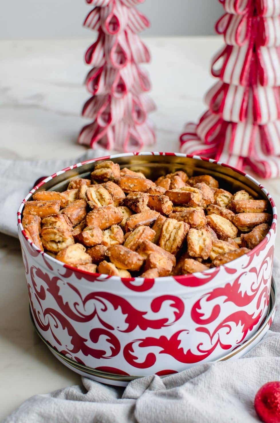 A red and white Christmas tin filled with Ranch Pretzel Bites with candy cane trees in the background. 