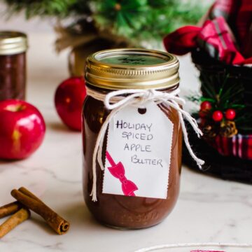 A jar of apple butter wrapped with twine and a gift tag with other jars and a Christmas tree in the background.